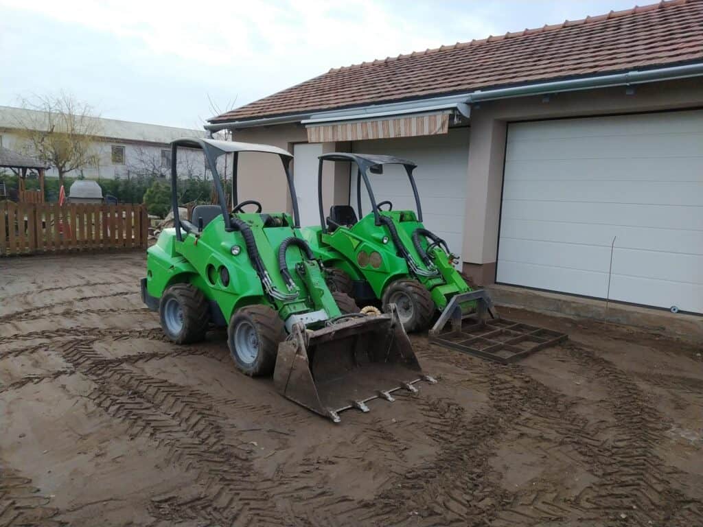 Két zöld gépi földmunka eszköz parkoló a ház előtt, a kert felújításához és tereprendezéshez.
