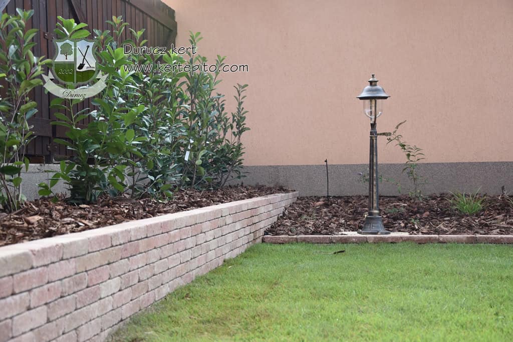 Kerti magaságyás építése téglából, zöld növényekkel és kültéri lámpással.