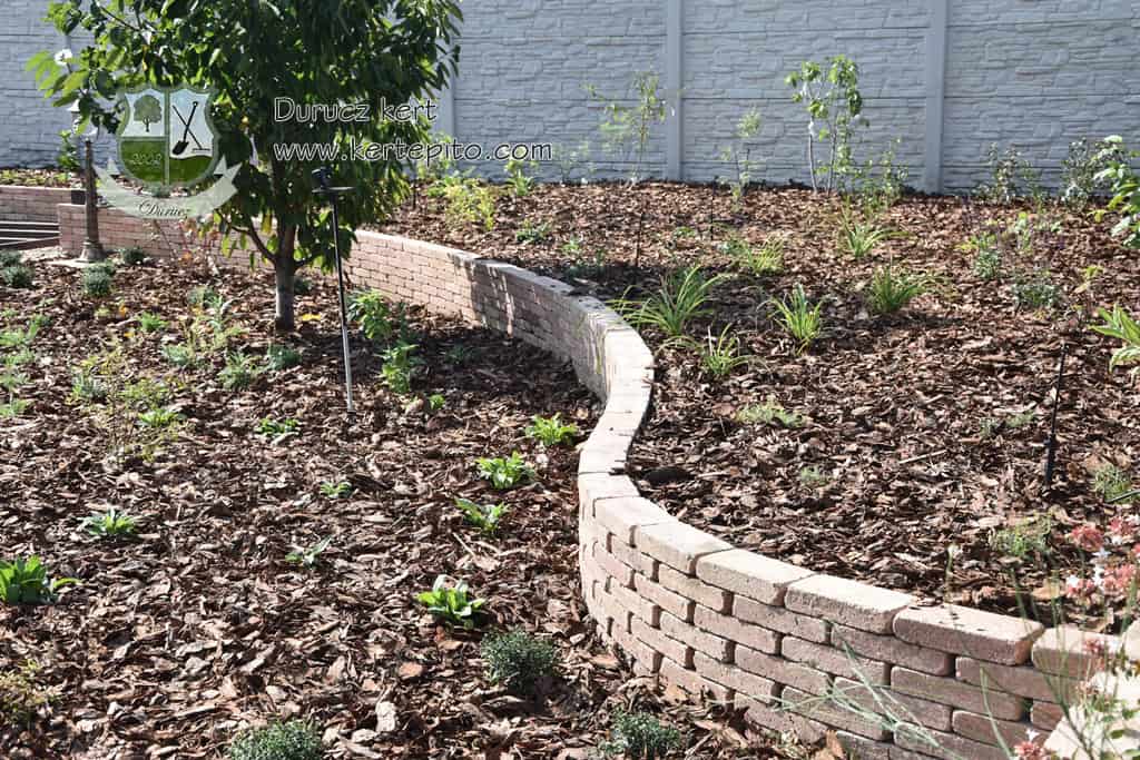 Támfal építés kerti területen, kőből és téglából, a növények védelmére és a terület határolására.